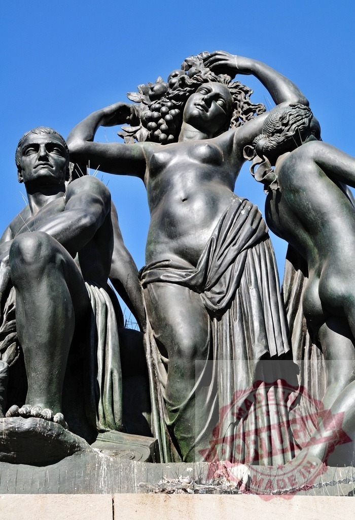 Bronze neoclassical sculpture depicting people with grapes against blue sky. Spain - Barcelona | Bronze sculpture featuring neoclassical figures with grapes, symbolizing abundance and harvest - Realized with Pictrs.com