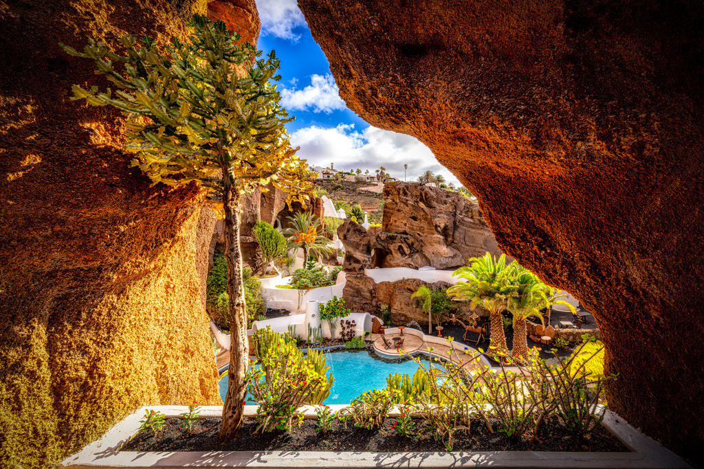 Blick in das von César Manrique konizpierte LagOmar Museum auf Lanzarote | Das Bild zeigt das LagOmar Museum, ein außergewöhnliches Anwesen in Nazaret auf der kanarischen Insel Lanzarote. Das Anwesen wurde in einen ehemaligen vulkanischen Steinbruch hineingebaut und ist berühmt für seine nahtlose Integration von Architektur in die natürliche Lavalandschaft. Details zum LagOmarArchitektur: Die Anlage wurde Anfang der 1970er Jahre von dem berühmten Inselkünstler César Manrique und dem Architekten Jesús Soto konzipiert. Sie besteht aus einem Labyrinth von Höhlen, Tunneln, Terrassen und einem zentralen Pool.Die Legende von Omar Sharif: Das Haus ist auch als "Casa Omar Sharif" bekannt. Der Legende nach kaufte der Hollywood-Schauspieler das Anwesen während Dreharbeiten auf der Insel, verlor es aber noch am selben Tag bei einem Bridge-Spiel gegen den Immobilienmakler Sam Benady. - Realisiert mit Pictrs.com