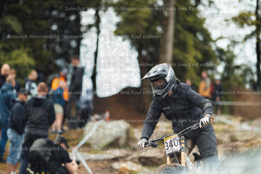 Mountainbike-Action im Wald | Das Bild zeigt einen Mountainbiker in voller Montur, der einen staubigen, unebenen Pfad durch einen bewaldeten Bereich hinunterfährt. Der Fahrer trägt einen schwarzen Helm mit Visier, eine dunkle Jacke und Handschuhe. Er trägt die Nummer 340 auf seinem Trikot. Im Hintergrund sind mehrere Zuschauer in verschiedenen Kleidungsstücken zu sehen, darunter einige mit orangefarbenen Warnwesten. Der Boden ist mit Staub bedeckt, der durch die Bewegung des Fahrers aufgewirbelt wird. Die Szene vermittelt ein Gefühl von Action und Abenteuer. - Realisiert mit Pictrs.com