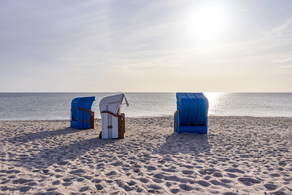 XXL Wandbild: Strandkörbe am Ostseestrand | Dieses XXL Wandbild im Querformat zeigt drei Strandkörbe am Ostseestrand in schöner Morgenstimmung. Über dem Meer ist die Sonne am fast wolkenlosen Himmel zu sehen. Durch die tiefstehende Sonne ergeben sich drei Schatten direkt an den Strandkörben. - Realisiert mit Pictrs.com