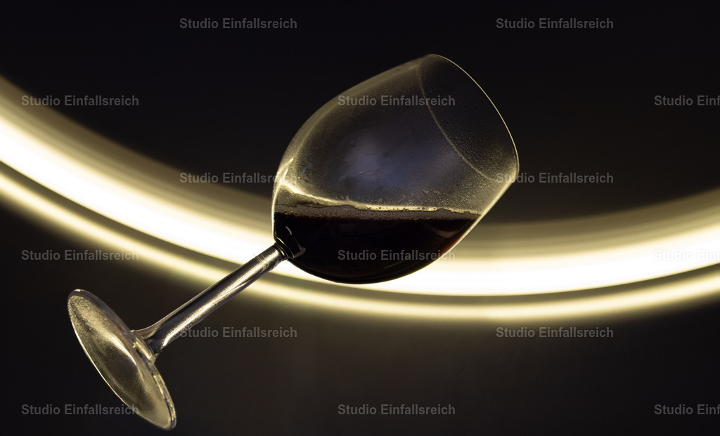 Dynamisches Weinglas mit Lichtspuren  | Eine kunstvolle High-Speed-Aufnahme eines schräg positionierten Weinglases, das mit einer dunklen Flüssigkeit gefüllt ist. Im Hintergrund erzeugen geschwungene, helle Lichtspuren (Light Painting) eine starke Dynamik und Tiefe. Die kühle, bläuliche Farbpalette verleiht dem Bild einen modernen, futuristischen Look. Ideal für exklusives Design, Bars oder moderne Wandkunst. - Realisiert mit Pictrs.com