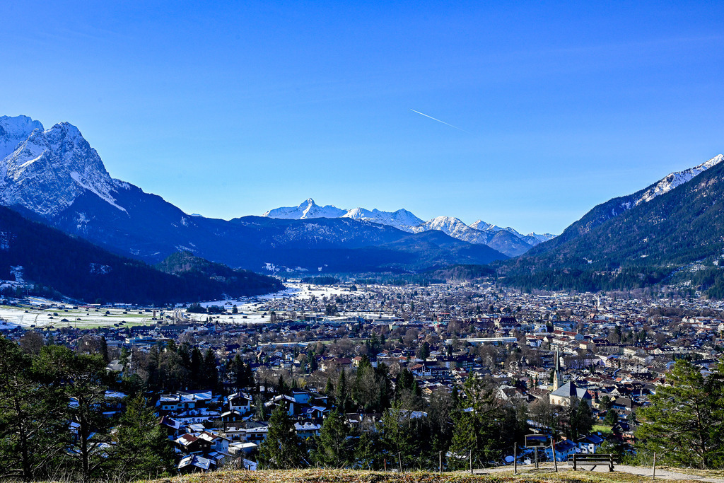Winterliches Talglück: Der Blick auf Garmisch-Partenkirchen | Ein friedlicher Zauber liegt über dem weiten Loisachtal: Sanft eingebettet in die mächtige Bergwelt der bayerischen Alpen ruht der malerische Ort Garmisch-Partenkirchen unter einem strahlend blauen Winterhimmel. Während im Tal die schneebedeckten Dächer und Wiesen im kühlen Schatten glitzern, thronen im Hintergrund die majestätischen, weiß gezuckerten Gipfel in hellem Sonnenlicht. Diese Aufnahme fängt die unendliche Weite, die erhabene Ruhe und die tiefe Sehnsucht nach einem klaren, winterlichen Tag in den Bergen perfekt ein. Ein Ort zum Innehalten und Durchatmen. - Realisiert mit Pictrs.com