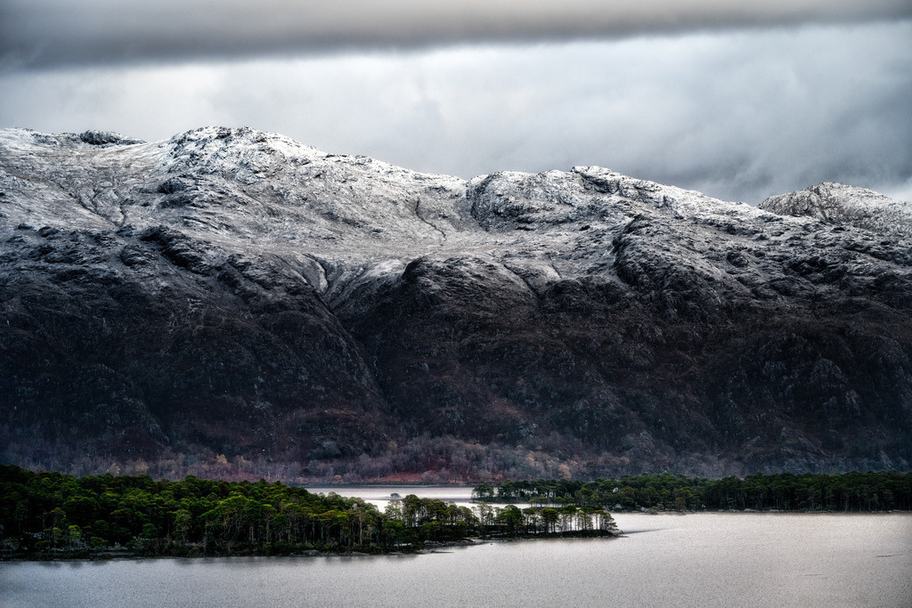Winterlandschaft am Loch Maree, Schottland | Eine dramatische Winterlandschaft zeigt schneebedeckte Berge unter einem bedeckten Himmel. Im Vordergrund erstreckt sich das dunkle Wasser des Loch Maree, unterbrochen von kleinen, bewaldeten Inseln. Die raue Textur der Bergflanken reicht von weissem Schnee bis zu dunklem Gestein und karger Vegetation. - Realisiert mit Pictrs.com