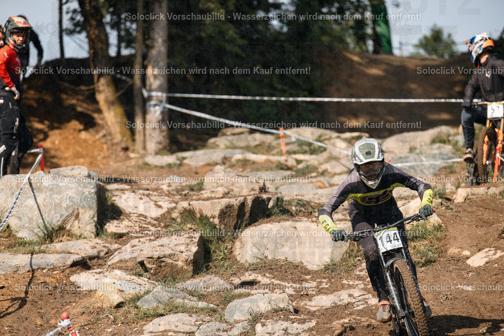 Mountainbike-Action | Das Bild zeigt einen Mountainbiker in voller Ausrüstung, der einen anspruchsvollen, steinigen Pfad hinunterfährt. Er trägt einen Helm, eine Schutzbrille, einen schwarzen Trikot mit gelben Akzenten und gelbe Handschuhe. Auf seinem Rücken befindet sich eine Nummer 44. Der Hintergrund besteht aus Felsen, Erde und vereinzelten Bäumen. Mehrere andere Mountainbiker sind im Hintergrund zu sehen. - Realisiert mit Pictrs.com