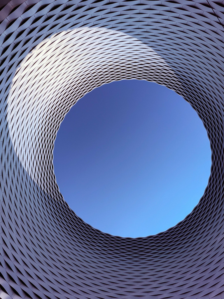 Wandbild Kunstdruck Poster – Fenster zum Himmel: Messe Basel im Sonnenlicht von Robert Steinke | Ein spektakuläres Meisterwerk der modernen Architektur-Fotografie, das die Grenzen zwischen Realität und Abstraktion verschwimmen lässt. Diese beeindruckende High-End-Aufnahme zeigt das weltberühmte "Fenster zum Himmel" der Messe Basel, entworfen von den visionären Star-Architekten Herzog &amp; de Meuron. Der Blick führt senkrecht nach oben durch den monumentalen, silbernen Aluminium-Trichter direkt in das makellose Blau des Himmels. In diesem einzigartigen Moment bricht sich das gleißende Sonnenlicht an der geschwungenen Wabenstruktur und lässt das Metall auf einer Seite magisch erglühen, während die andere Seite in elegantem Schatten verweilt. Eine faszinierende Dynamik aus Licht, Geometrie und unendlicher Weite.Dieses exklusive und minimalistische Kunstwerk ist der perfekte, repräsentative Blickfang für ein stilvolles Büro, das moderne Wohnzimmer, ein helles Design-Loft oder den repräsentativen Eingangsbereich.Um den metallischen Glanz der Wabenstruktur und das strahlende Blau des Himmels-Zentrums mit brachialer Tiefenwirkung an deine Wand zu bringen, empfehle ich dringend den Druck auf unseren High-End-Premium-Materialien: Erlebe kinoreife Leuchtkraft auf edlem Acrylglas oder eine hochmoderne, absolut edle und reflexionsfreie Galerie-Optik auf robustem Alu-Dibond. Verwandle deine Räume in eine moderne Kunstgalerie! - Realisiert mit Pictrs.com