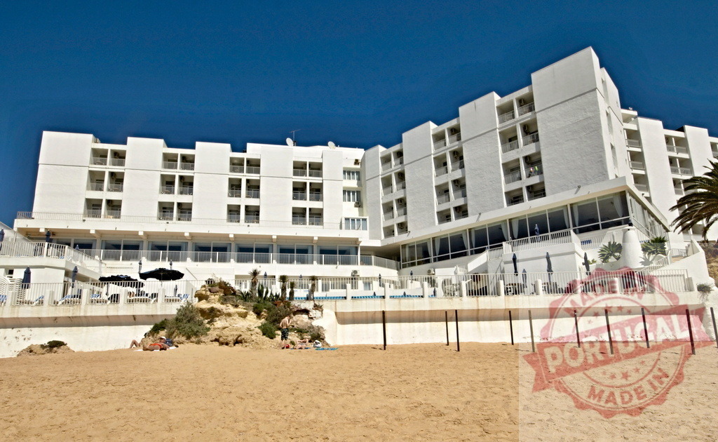 Hotel building with balconies overlooking sandy beach | White modern hotel building featuring balconies and terraces on a sandy beach under blue sky - Realized with Pictrs.com