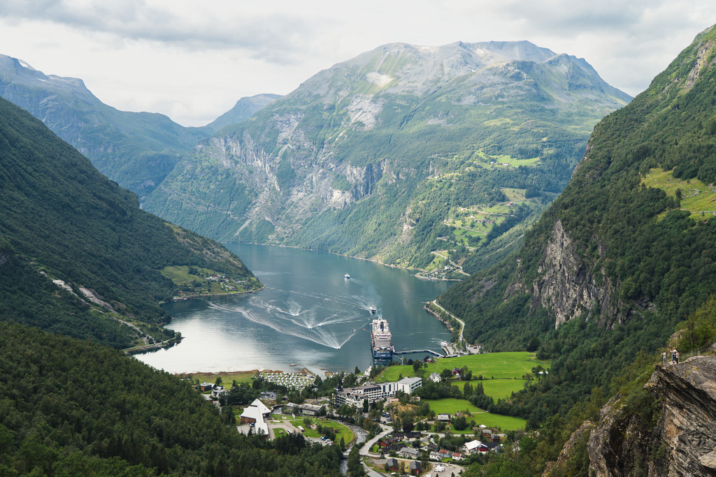 Mein Schiff 4 im Geirangerfjord | Ein Blick von den Höhenzügen auf den Geirangerfjord, einen der bekanntesten Fjorde Norwegens (UNESCO-Welterbe). Die Aufnahme zeigt die Mein Schiff 4 am Liegeplatz vor dem Ort Geiranger, umgeben von steilen Felswänden und der typischen skandinavischen Fjordlandschaft. Das Bild dokumentiert die beeindruckenden Größenverhältnisse zwischen moderner Kreuzfahrtschifffahrt und der unberührten Natur der norwegischen Westküste. - Realisiert mit Pictrs.com
