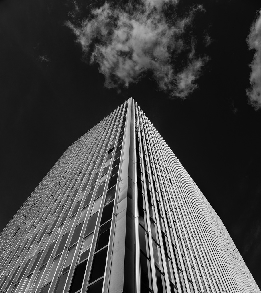 Into the Sky | This fine art architectural photograph transforms a modern high-rise into a bold geometric statement. Shot from a dramatic low angle, the building rises sharply into the dark sky, while the cloud above adds tension and atmosphere. The monochrome treatment emphasizes structure, rhythm and contrast, making this piece ideal for contemporary interiors, offices and minimalist spaces. - Realisiert mit Pictrs.com
