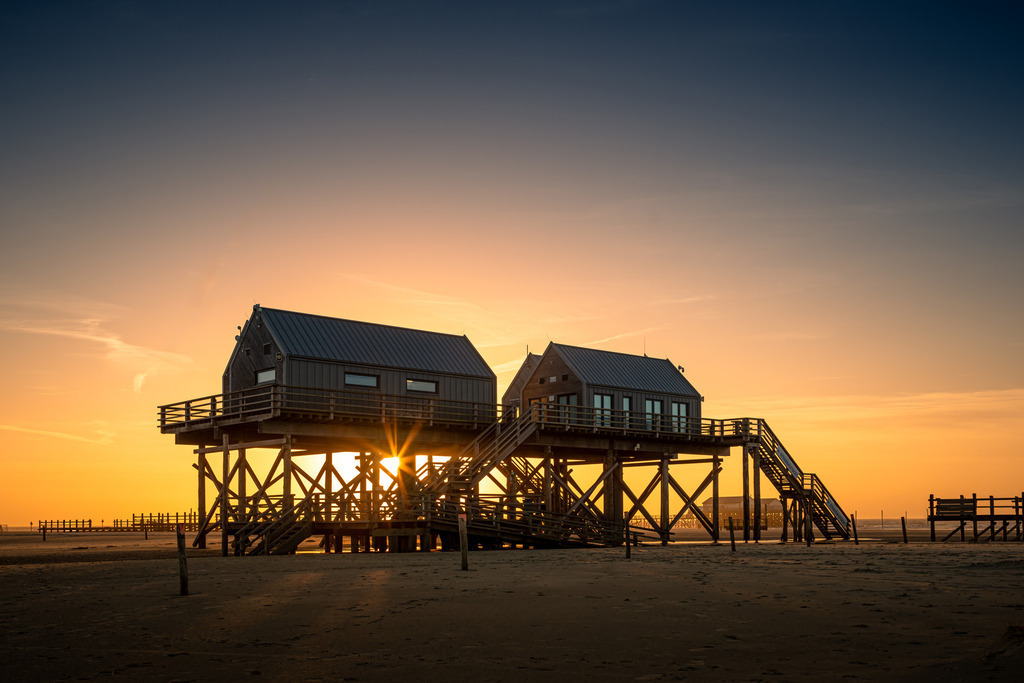 Sonnenuntergang in St. Peter Ording | Auf dem Bild sind die charakteristischen Pfahlbauten von St. Peter-Ording bei Sonnenuntergang zu sehen. Die Holzhäuser stehen auf hohen Stelzen, um sie vor Hochwasser zu schützen. Sie sind über Holztreppen und -plattformen zugänglich und bieten einen eindrucksvollen Blick auf das Meer und den Strand. Die warmen Sonnenstrahlen tauchen die Szene in ein stimmungsvolles, goldenes Licht.