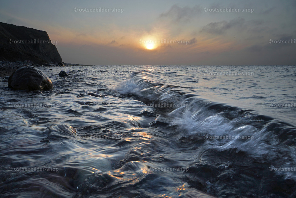 Mystische Stimmung am Ostseeufer | Eine Welle rollt dem Steinestrand vor der Steilküste entgegen. Der Abendhimmel ist leicht verhangen und das Sonnenlicht etwas fahl.