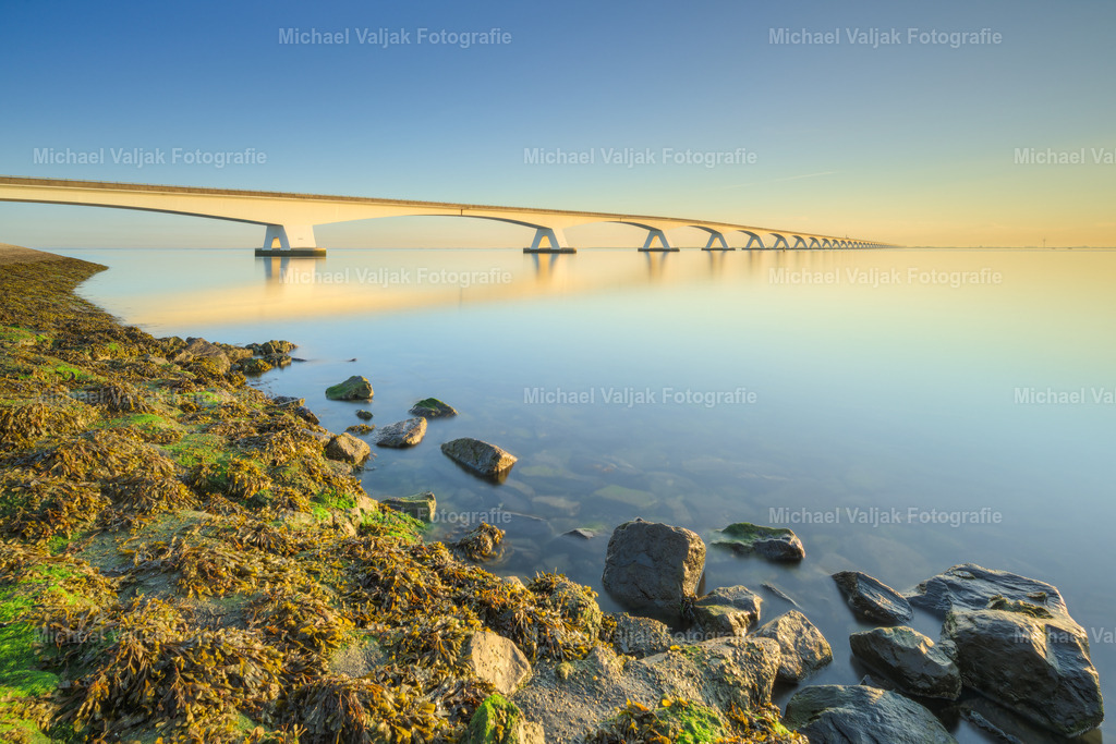 Morgens an der Zeelandbrücke | Die Zeelandbrücke liegt im ersten warmen Licht des Morgens und erstreckt sich eindrucksvoll über die stille Weite des Wassers. Im Vordergrund zeichnen sich die Steine am Ufer deutlich ab und bilden einen natürlichen Rahmen für die weit in die Ferne führende Brücke.Die Langzeitbelichtung verwandelt die Wasseroberfläche in eine sanfte, ruhige Fläche und verstärkt damit die friedliche Stimmung dieses frühen Morgens. Das goldene Licht der gerade aufgegangenen Sonne lässt die Brücke warm aufleuchten und setzt einen eindrucksvollen Kontrast zum kühlen Morgenhimmel.Ein ruhiges und atmosphärisches Landschaftsfoto aus Zeeland, Niederlande, das die besondere Stimmung der ersten Sonnenstrahlen an der Küste einfängt. - Realisiert mit Pictrs.com