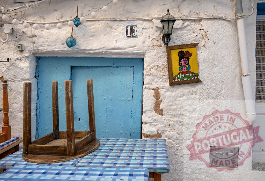 Rustic cafe table with blue checkered tablecloth and vibrant painting | Rustic cafe table with an inverted stool, blue door, and colorful painting on a whitewashed wall - Realisiert mit Pictrs.com