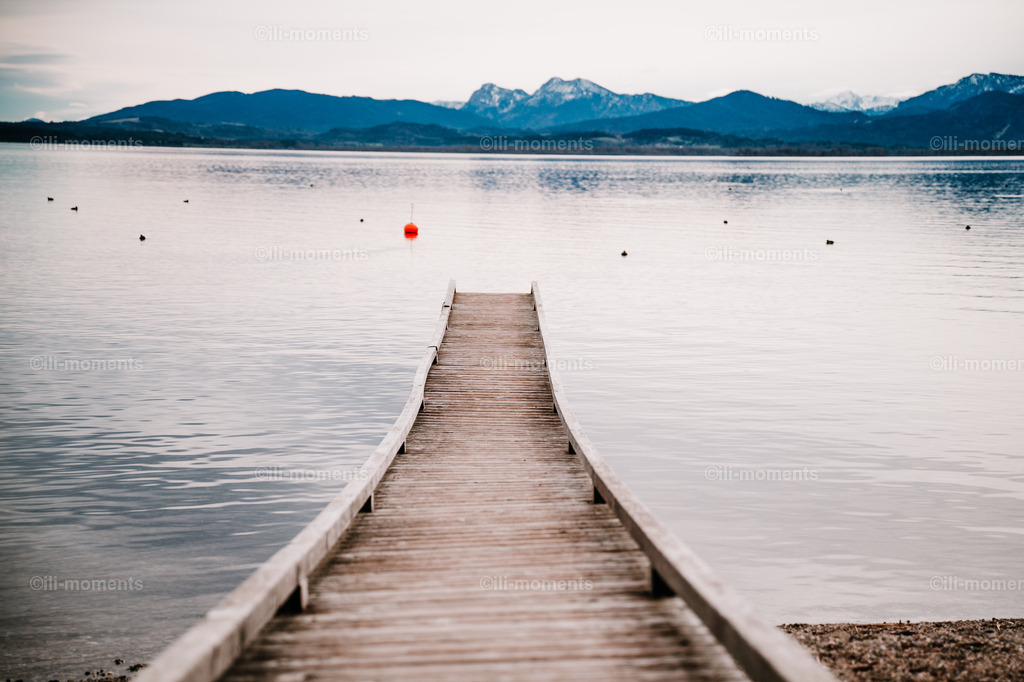 Weg zum See | Ein ruhiger Steg führt in einen stillen See, dahinter erheben sich majestätisch die Berge – ein stimmungsvolles Fotokunstwerk voller Weite und Sehnsucht. Ideal als Kunstdruck zur Gestaltung ruhiger, inspirierender Räume. - Realisiert mit Pictrs.com