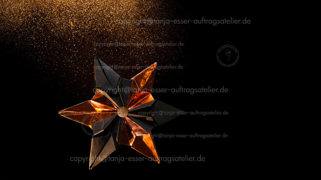 Star from black cardboard and bronze colored foil in front of black background | Feierlicher 6 zackiger Papierstern und fliegendes Glitzerpartikel vor schwarzem Hintergrund. Rötlich leuchtendes kupferfarbiges Folienpapier und matt schwarzer Fotokarton.  Copy Space. 