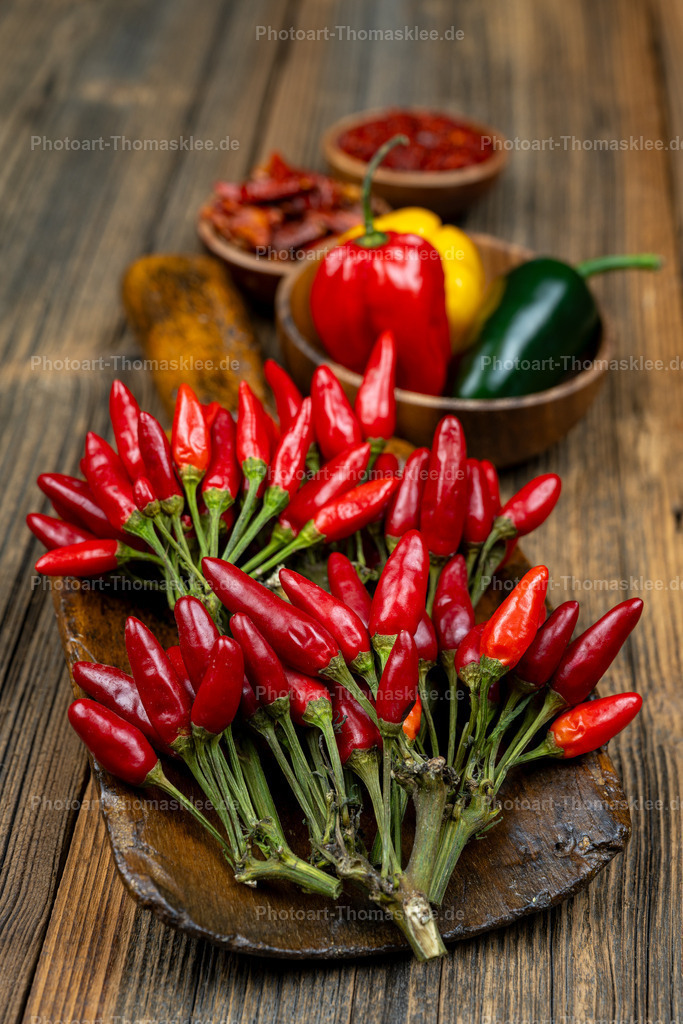 Feurige rote Chilischoten auf rustikalem Holztisch | Auf einem warmen Holztisch präsentieren sich leuchtend rote Chilischoten in natürlicher Schönheit. Die kleinen scharfen Schoten mit grünen Stielen liegen gebündelt auf einer dunklen Holzschale, während im Hintergrund bunte Paprika und weitere Gewürze in rustikalen Schälchen zu sehen sind. Die Holzmaserung des Tisches unterstreicht den authentischen Charakter dieser Küchenszene. Diese harmonische Anordnung zeigt die Vielfalt scharfer Gewürze und lädt zum Kochen mit intensiven Aromen ein. - Realisiert mit Pictrs.com