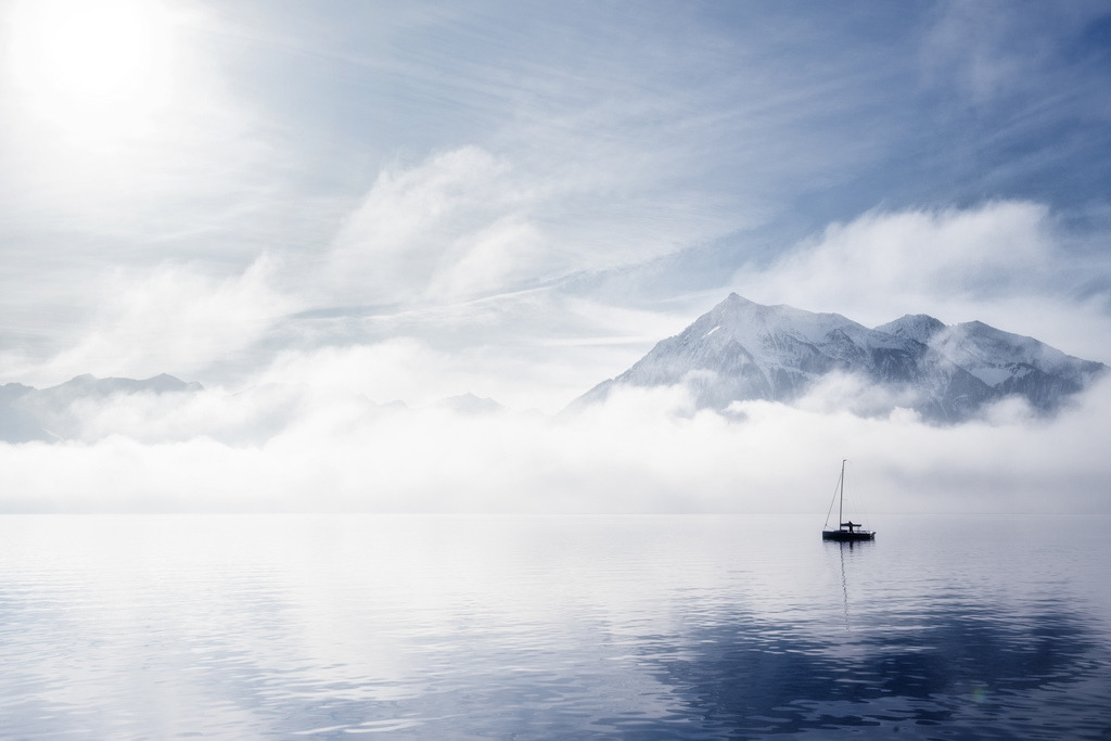 Nebelkapitän | Ein Segelboot gleitet ruhig auf dem spiegelglatten Wasser des Thunersees, umgeben von dichtem Nebel, der die Basis der schneebedeckten Berge verhüllt. Im Hintergrund erhebt sich der markante Niesen, dessen Gipfel aus den Wolken ragt, während die Wintersonne durch eine helle Wolkendecke bricht und die Szene in kühles Licht taucht. - Realisiert mit Pictrs.com
