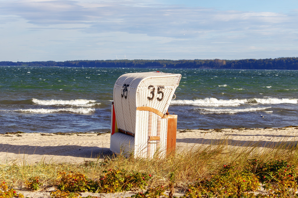Wandbild: Strandkorb am Meer in Eckernförde  | Dieses Wandbild zeigt einen einzelnen weißen Strandkorb am Sandstrand von Eckernförde an der Ostsee. Der Korb steht ruhig im Vordergrund, umgeben von etwas Strandvegetation. Dahinter breitet sich das Meer aus – bewegt, lebendig, mit einer bewaldeten Küstenlinie am Horizont. Der Himmel ist leicht bewölkt und öffnet die Szene nach oben. Die Komposition lebt von der Ruhe des Moments, der Klarheit der Formen und dem Gefühl von Rückzug und Weite. Ideal als Wandbild für naturnahe Wohnkonzepte – ob als Leinwandbild, Acrylglasbild, Alu-Dibond FineArt Print oder als Akustikbild. Ein stiller, maritimer Akzent für Wohnzimmer, Büro oder Ferienwohnung. - Realisiert mit Pictrs.com