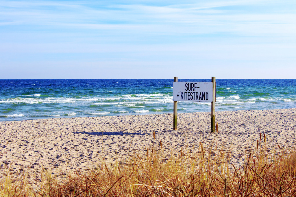 Wandbild: Surf- und Kitestrand in Weidefeld | Dieses Wandbild im Querformat bringt den maritimen Flair von Weidefeld direkt in Ihr Zuhause. Der Sandstrand wird durch das Schild "Surf-Kitestrand" belebt und der Vordergrund ist mit Strandhafer geschmückt. Ein klarer blauer Himmel mit zarten Schleierwolken vervollständigt diese idyllische Küstenlandschaft. - Realisiert mit Pictrs.com