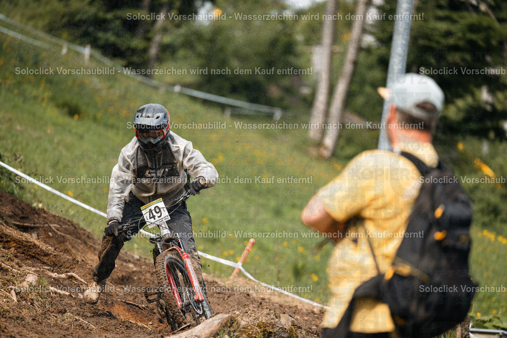 Mountainbike-Abenteuer | Das Foto zeigt einen Mountainbiker in voller Ausrüstung, der einen steilen, grasbewachsenen Hang hinunterfährt. Er trägt einen weißen Helm mit einer schwarzen Visier und eine passende Schutzkleidung. Er trägt die Nummer 49 auf seinem Oberkörper. Ein Zuschauer steht am Rande des Weges und beobachtet ihn. Der Hintergrund besteht aus Bäumen und einem bewölkten Himmel. - Realisiert mit Pictrs.com