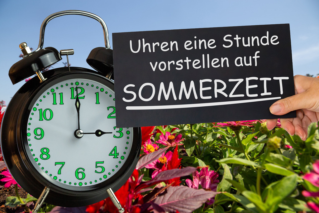 Uhren eine Stunde vorstellen auf Sommerzeit | (Fotomontage) Zeitumstellung Sommerzeit Symbolbild: Wecker auf einer Blumenwiese und Schild mit Text UHREN EINE STUNDE VORSTELLEN AUF SOMMERZEIT - Realisiert mit Pictrs.com