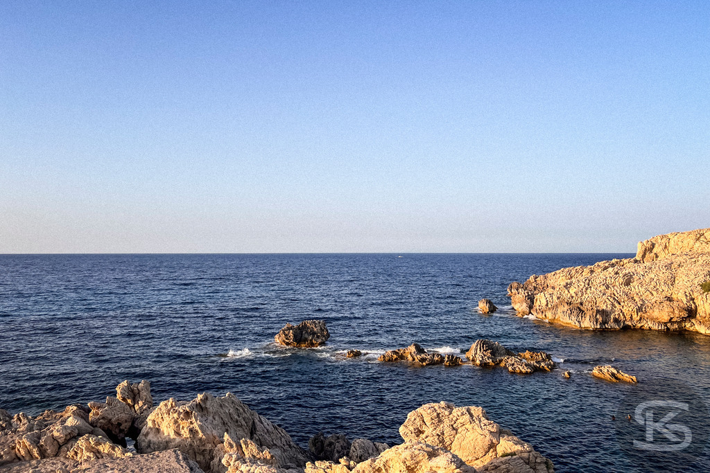 Abendruhe an der Cala Agulla – Exklusives Wandbild der mallorquinischen Felsenküste | Küstenfelsen im ruhigen Wasser der Cala Agulla auf Mallorca während der Abenddämmerung mit Blick auf die entfernte Silhouette der Küstenlinie. - Realisiert mit Pictrs.com