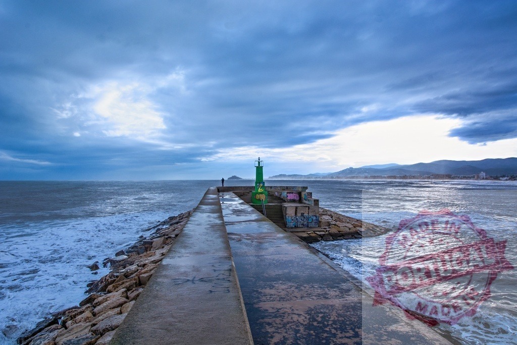 Man walking on pier under cloudy sky, facing rough sea and distant beach | Concrete pier with green beacon under cloudy sky, man walking towards the sea with waves crashing, beach and mountains in the background - Realized with Pictrs.com