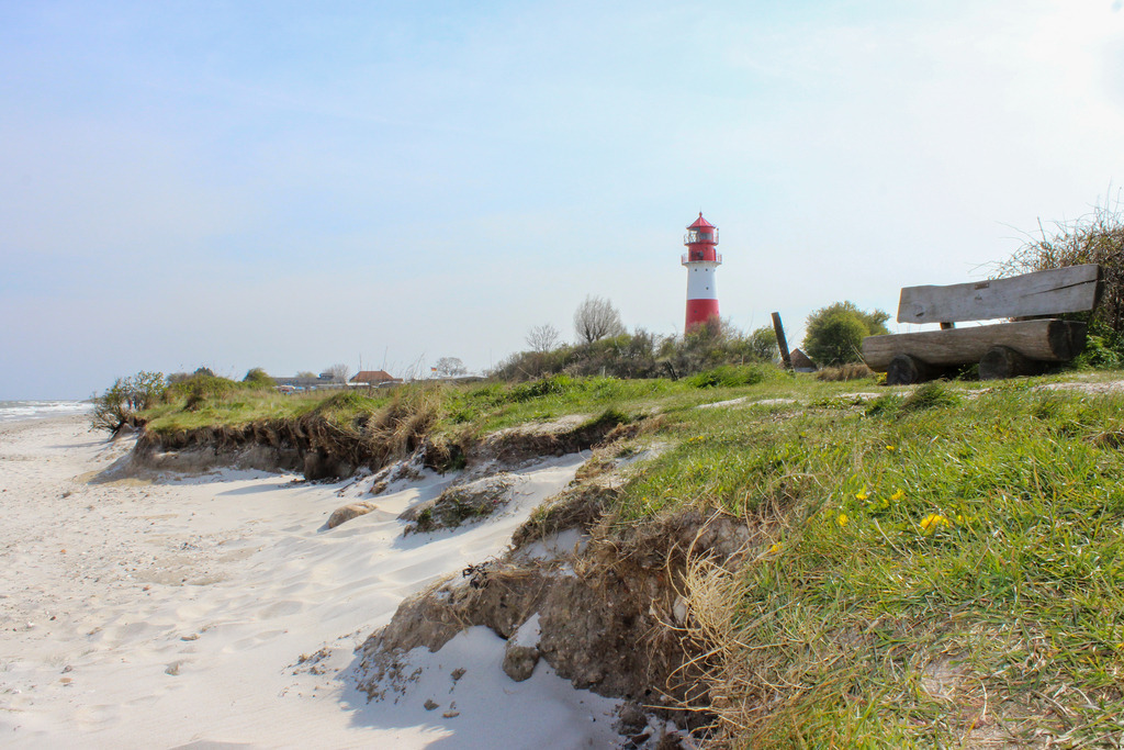 Wandbild: Leuchtturm in Falshöft im Frühling | Dieses Wandbild im Querformat zeigt den Strand in Falshöft im Frühling. In der Ferne kann man den Leuchtturm gut erkennen. Auf der rechten Seite befindet sich eine Bank zum verweilen. Der blaue Himmel ist fast wolkenlos.  - Realisiert mit Pictrs.com