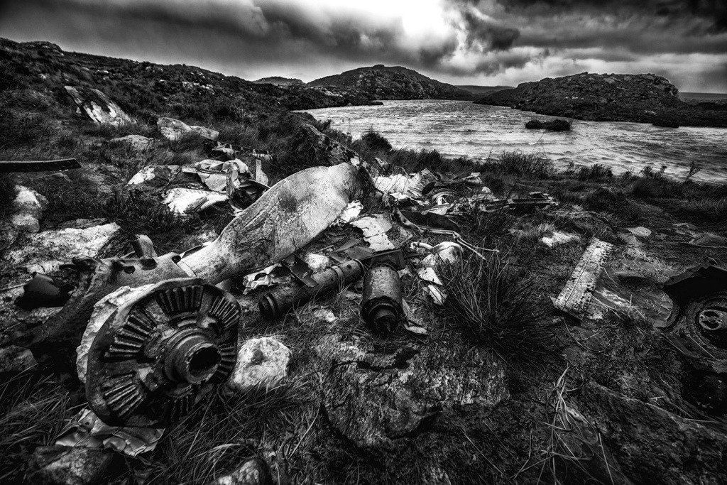 Absturzstelle eines USAAF B-24H Liberator-Bombers | Am 13. Juni 1945 stürzte ein USAAF B-24H Liberator nahe Gairloch, Schottland, ab – alle 15 Besatzungsmitglieder starben. Das Flugzeug war von Prestwick in die USA unterwegs, verliess den Kurs vermutlich wegen Navigationsfehler, Technik oder Wetter und stürzte beim Versuch einer Notlandung im Loch Gairloch in die Hügel oberhalb der Fairy Lochs ab, nachdem es den Berg Slioch streifte. Die Trümmer liegen heute verstreut am Ufer und auf dem Hügel; einige Teile wie Rumpf, Propeller und Triebwerke sind noch erkennbar. Der Ort ist eine Kriegsgräberstätte mit Gedenktafel und Besucher-Gedenkobjekten.Verstreute Wrackteile eines Flugzeugs liegen im Vordergrund auf felsigem, grasbewachsenem Gelände. Dahinter erstreckt sich ein windgepeitschter See, umgeben von hügeliger Landschaft unter einem dramatischen, bewölkten Himmel. Die Schwarz-Weiss-Darstellung betont die raue Textur der Überreste und die melancholische Atmosphäre der Szenerie. - Realisiert mit Pictrs.com