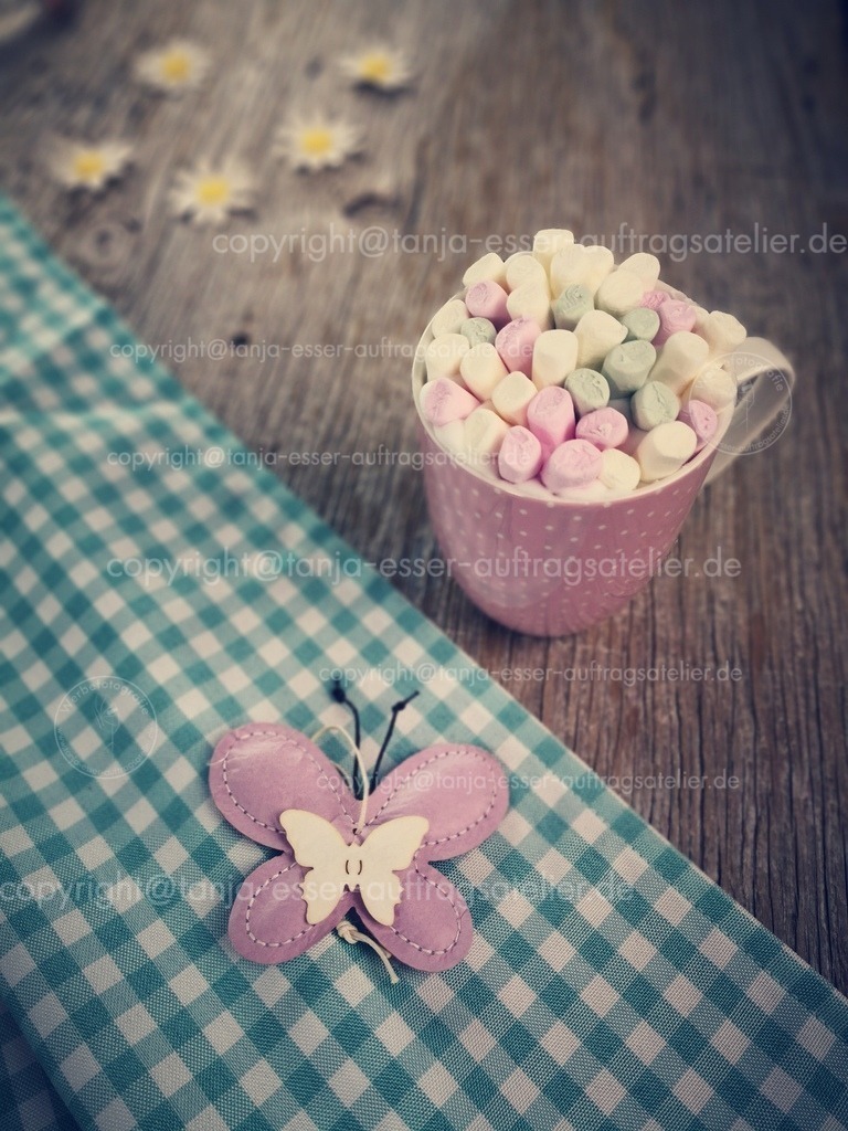 Frühling Tasse Kakao mit Marshmallows D | Ein rosa Becher mit heißem Kakao. Oben drauf schwimmen viele pastellfarbige kleine Marshmallow. Im Vordergrund ist eine karierte Tischdecke mit einem Schmetterling. 