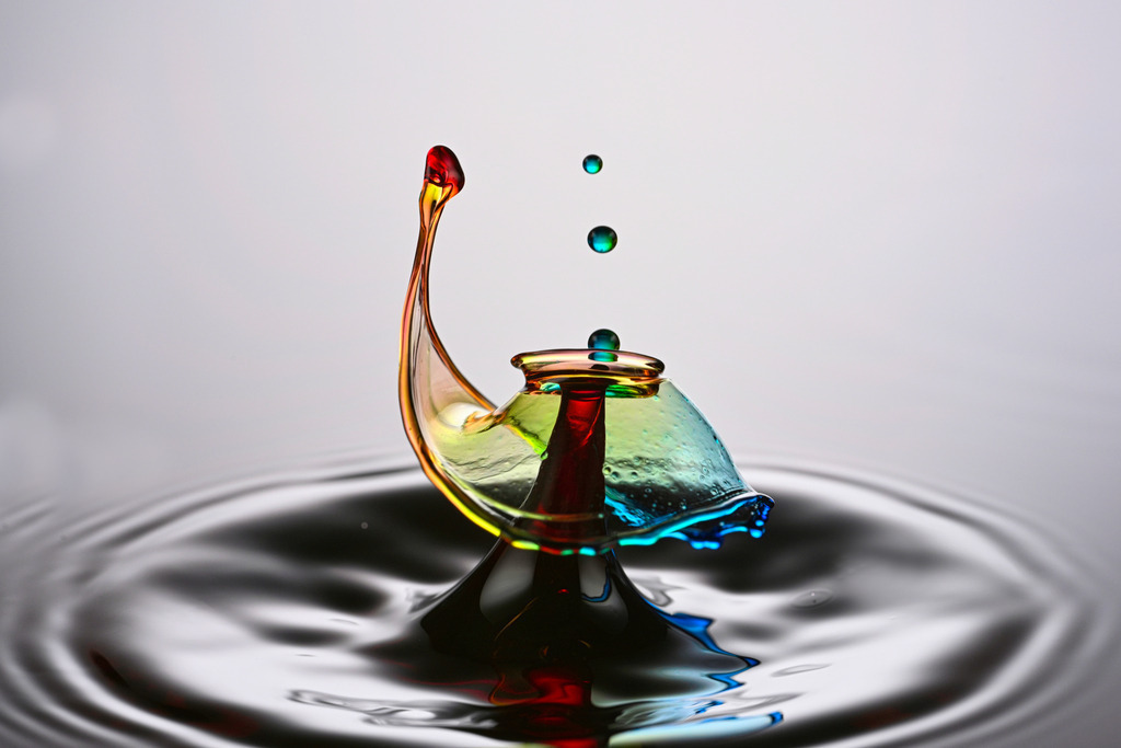 Farbenfroher Tropfen | Eine farbenfrohe Wassertropfen-Kollision in Nahaufnahme. Die Aufnahme zeigt den Moment, in dem zwei Tropfen aufeinandertreffen und eine elegante, kronenförmige Wasserstruktur mit Regenbogenfarben bilden. Die glatte Wasseroberfläche reflektiert sanft die Farben – ein faszinierendes Beispiel für Highspeed-Fotografie und Flüssigkeitskunst.