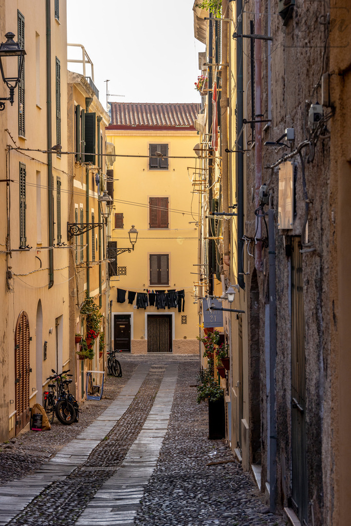 Quiet Alleyways | In the golden morning light, the narrow cobbled street of Alghero tells stories of everyday life — bicycles leaning on walls, laundry swaying gently in the breeze, and a calm that belongs only to old towns by the sea. - Realisiert mit Pictrs.com