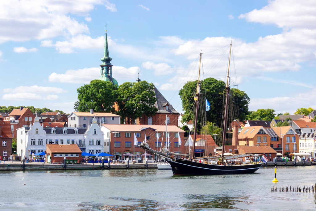 Wandbild: Traditionssegler auf der Schlei in Kappeln | Dieses Wandbild im Querformat zeigt einen Traditionssegler auf der Schlei in Kappeln. Auf der anderen Schleiseite ist der Hafen in Kappeln zu sehen. Am blauen Himmel befinden sich schöne sommerliche Wolken.  - Realisiert mit Pictrs.com