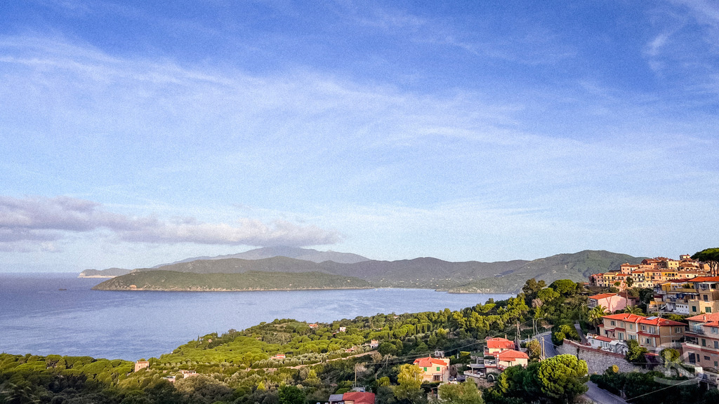 Morgenerwachen über Capoliveri: Panorama-Blick auf Elba – Wandbild | Weitwinkel-Ansicht von Capoliveri auf Elba bei Sonnenaufgang; rechts im Bild die terrassenförmig angelegten Häuser des Dorfes, links der Blick über die bewaldete Küste und das blaue Meer bis zum Horizont. - Realisiert mit Pictrs.com