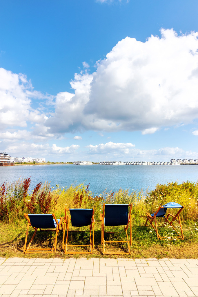 Wandbild: Liegestühle an der Promenade in Olpenitz  | Dieses Wandbild im Hochformat zeigt Liegestühle an der Promenade in Olpenitz. In der Ferne sind die Ferienwohnungen am Meer zu sehen. Am blauen Himmel befinden sich einige helle Wolken.  - Realisiert mit Pictrs.com