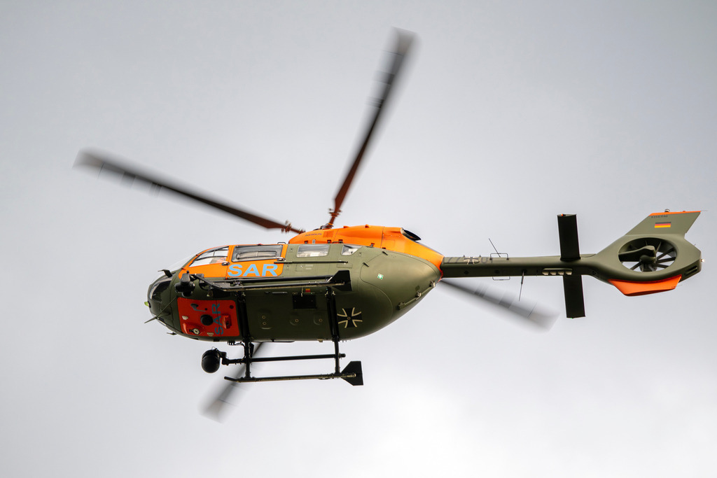 SAR | Das Foto zeigt einen Hubschrauber der deutschen Bundeswehr im Flug gegen einen bewölkten, hellgrauen Himmel. Es handelt sich um ein Modell vom Typ Airbus H145 (LUH SAR) in militärischer Tarnlackierung (Olivgrün) mit auffälligen, leuchtend orangefarbenen Markierungen und dem Schriftzug „SAR“ (Search and Rescue). Das Eiserne Kreuz als Hoheitszeichen ist deutlich auf der Unterseite und an den Seiten erkennbar. Die Rotorblätter sind durch die Bewegung leicht unscharf dargestellt, was die Dynamik des Fluges unterstreicht. - Realisiert mit Pictrs.com