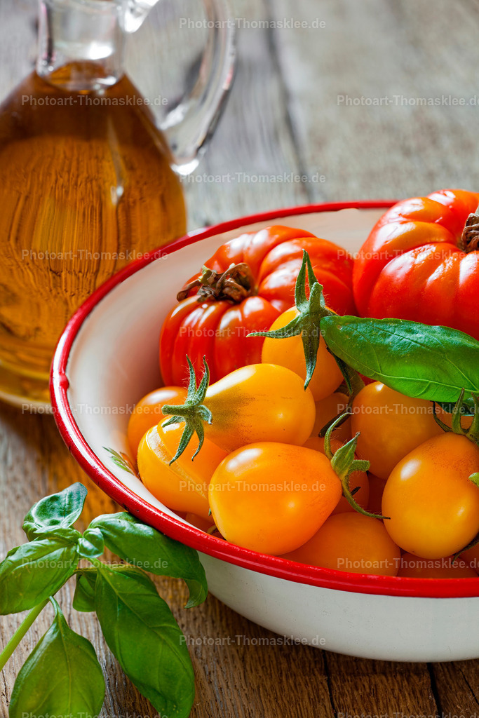 Bio Tomaten in Emailleschale, Basilikum und Olivenöl | Saftige Tomaten verschiedener Sorten füllen eine nostalgische Emailleschüssel und schaffen eine authentische Küchenatmosphäre. Die reifen Früchte zeigen ihre natürliche Schönheit mit glänzender Haut und frischen grünen Kelchblättern. Eine kleine Olivenölkaraffe aus klarem Glas und einige Basilikumblätter komplettiert diese klassische Zutatenkombination. Das warme Holz im Hintergrund unterstreicht den ländlichen Charme dieser Foodfotografie und weckt Appetit auf mediterrane Gerichte. - Realisiert mit Pictrs.com