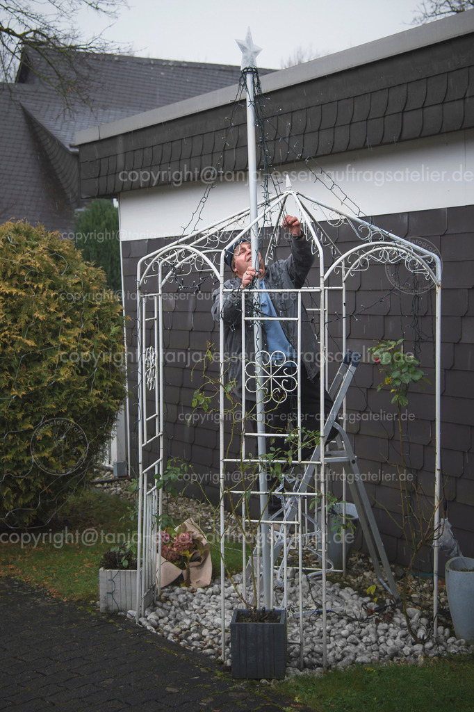 Advent season and Christmas - the chain of lights are installed in the garden, even if it rains | Adventszeit. Ein Mann installiert in der regnerischen Novemberzeit eine Lichterkette an einem Metall Pavillon für Blumen im Vorgarten. Er steht auf einer Leiter und trägt eine Mütze. 