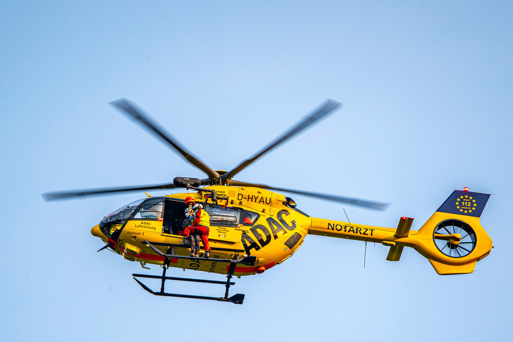 Bergretter | Ein markanter gelber ADAC-Rettungshubschrauber (Christoph 1) befindet sich im Schwebeflug vor einem klaren, hellen Himmel. An der Seite ist die Rettungswinde im Einsatz: Ein Luftretter sichert eine Person, während sie gemeinsam am Seil unterhalb der offenen Kabinentür hängen – ein intensiver Einblick in die lebensrettende Arbeit der Bergwacht oder Luftrettung. - Realisiert mit Pictrs.com