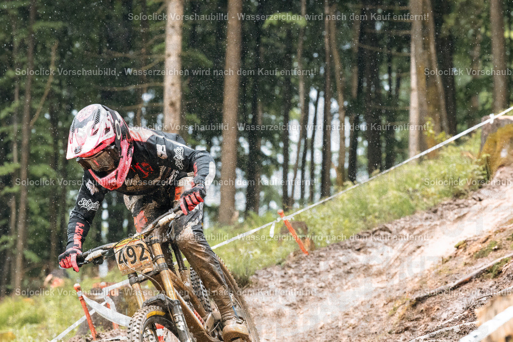 Adrenalin auf zwei Rädern | Ein Mountainbiker in voller Ausrüstung rast einen staubigen, schmalen Trail in einem dichten Wald hinunter. Er trägt einen roten Helm mit einem auffälligen Muster, eine passende Schutzkleidung und Handschuhe. Das Gelände ist steil und mit Wurzeln und Steinen übersät. Der Fahrer scheint sich voll und ganz auf die Fahrt zu konzentrieren und meistert die Herausforderungen des Trails mit Geschick und Präzision. - Realisiert mit Pictrs.com