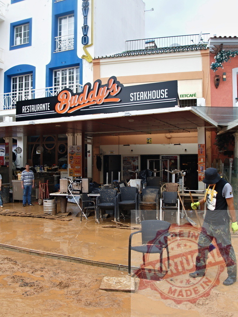 People cleaning restaurant Buddy's Steakhouse after flood | Albufeira Flood - discruction by weather problems Workers removing mud and damaged furniture from a restaurant after natural disaster - Realized with Pictrs.com