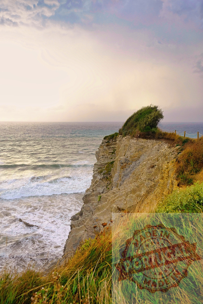 Rocky cliff overlooking ocean waves under dramatic sky | Rocky cliff edge with green grass overlooking crashing ocean waves under a cloudy sky - Realized with Pictrs.com