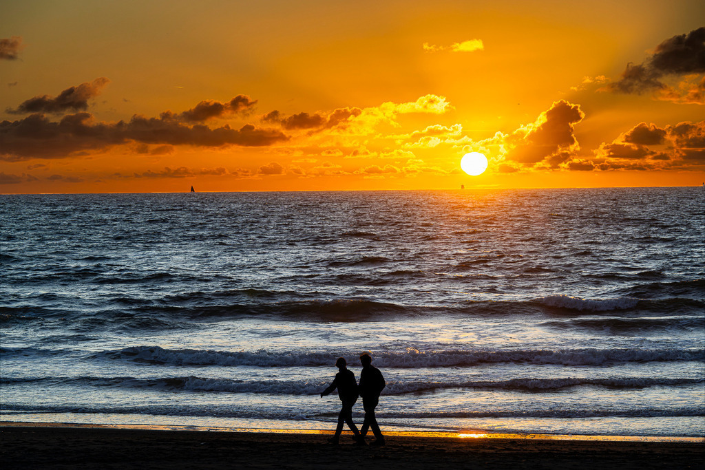 Paar genießt die Abendstimmung am Strand | Zwei Spaziergänger zeichnen sich als dunkle Silhouetten am Sandstrand der niederländischen Insel Texel vor der Kulisse der Nordsee ab. Die tief stehende Sonne taucht den Himmel und die sanften Wellen in intensives, warmes Orange-Gelb. Am weit entfernten Horizont ist die Silhouette eines Segelboots zu erkennen. Ein stimmungsvolles Motiv für redaktionelle Berichte über Tourismus, Strandurlaub, Romantik, Erholung an der Küste und Naturerlebnisse. - Realisiert mit Pictrs.com