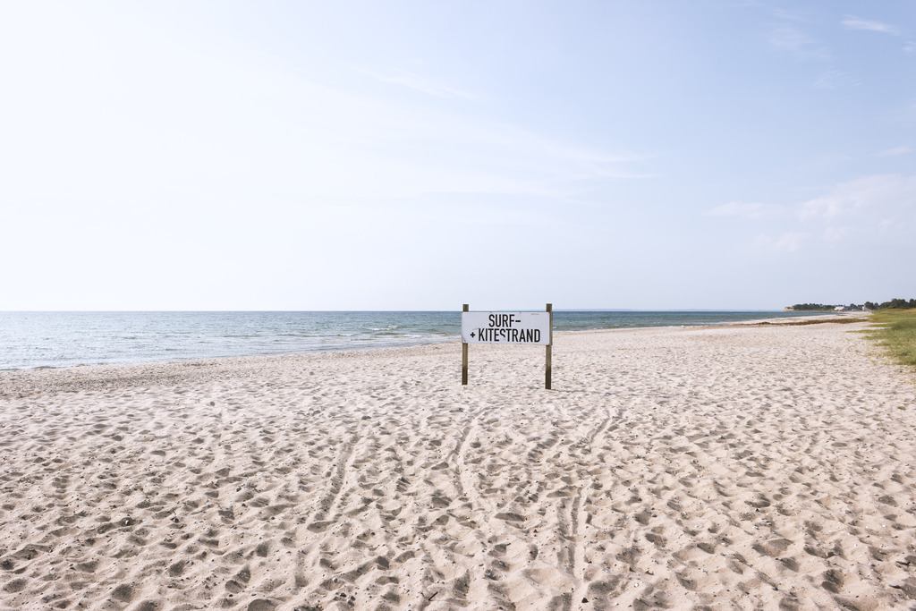Wandbild: Weite am Weidefelder Strand | Der Weidefelder Strand zeigt sich hier in seiner offenen, weiten Schönheit: heller Sand, ruhige Ostsee und ein klarer Himmel, der sich weit über die Küste spannt. Das Schild zum Surf‑ und Kitestand steht wie ein stiller Hinweis auf die Freiheit, die dieser Strand ausstrahlt — Raum für Wind, Wasser und die Leichtigkeit des Augenblicks. Die sanften Wellen und die unberührte Weite schaffen eine Atmosphäre, die zugleich klar, ruhig und inspirierend wirkt. Als Wandbild bringt diese Szene die helle, weite Küstenlandschaft direkt in den Raum und schafft einen Moment von maritimer Gelassenheit, der lange nachwirkt.