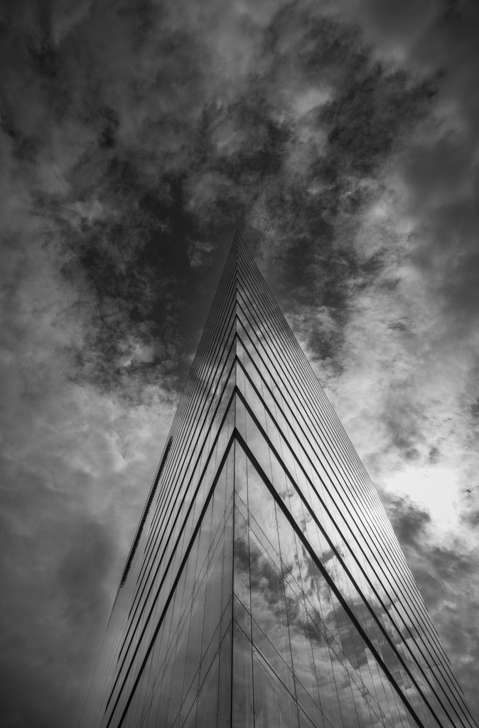 The Edge | Die markante Dreiecksfassade eines modernen Hochhauses in Düsseldorf ragt wie eine Klinge in den dramatischen Himmel. Die Spiegelungen in der Glasfassade und die kontrastreiche Schwarzweißbearbeitung betonen Geometrie, Perspektive und Architektur. Ein minimalistisches Fine-Art-Motiv, das die Dynamik moderner Stadtarchitektur eindrucksvoll einfängt. - Realisiert mit Pictrs.com