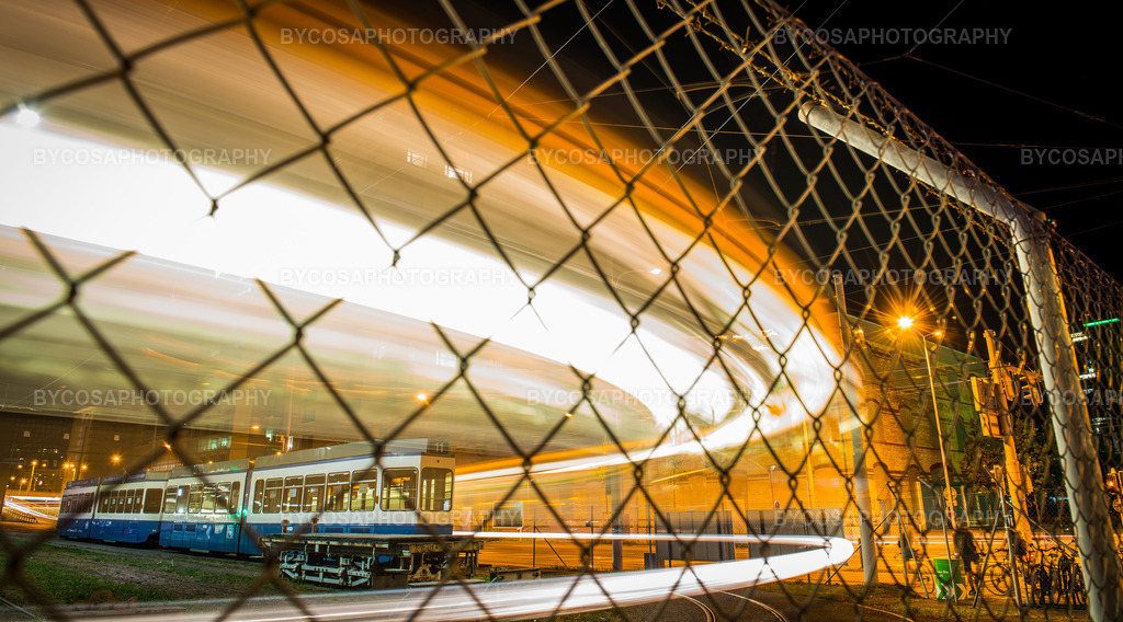 Urban Motion _ Zürich Tram Light Trails | Eine ausdrucksstarke FineArt-Fotografie, die die pure Energie Zürichs bei Nacht einfängt. Durch den Metallzaun saust eine Straßenbahn in einem langbelichteten Bogen vorbei und erzeugt faszinierende Lichtspuren, die eine leuchtende, fast abstrakte Schleife bilden.Die Kombination aus industriellen Elementen, Bewegungsunschärfe und Nachtlicht macht diesen Druck zu einem einzigartigen urbanen Kunstwerk – perfekt für moderne Räume, Büros, Bars und alle Liebhaber dynamischer, abstrakter Fotografie. - Realisiert mit Pictrs.com