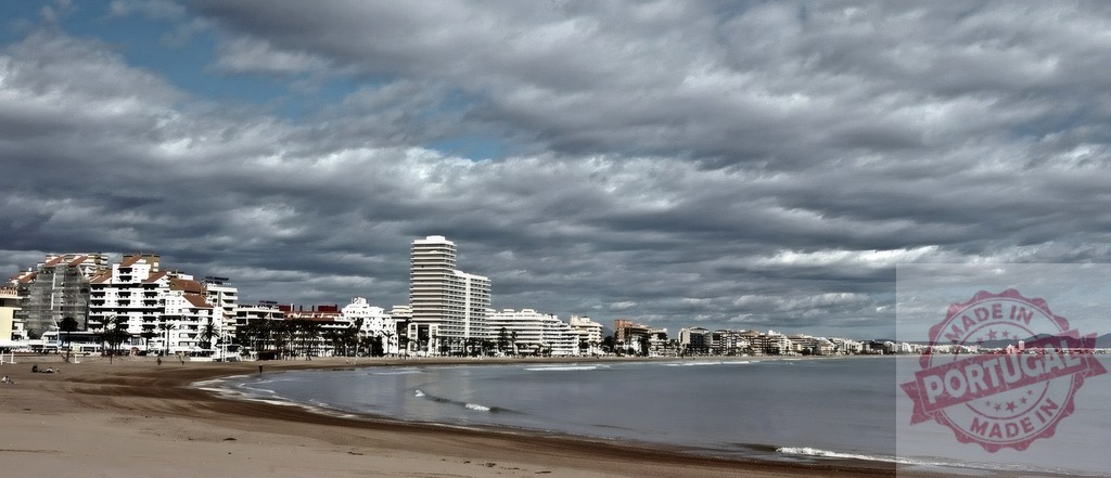 City beach with buildings along the coastline under cloudy sky | Urban beach front with apartment buildings, a sandy shore, and calm sea under an overcast sky - Realized with Pictrs.com