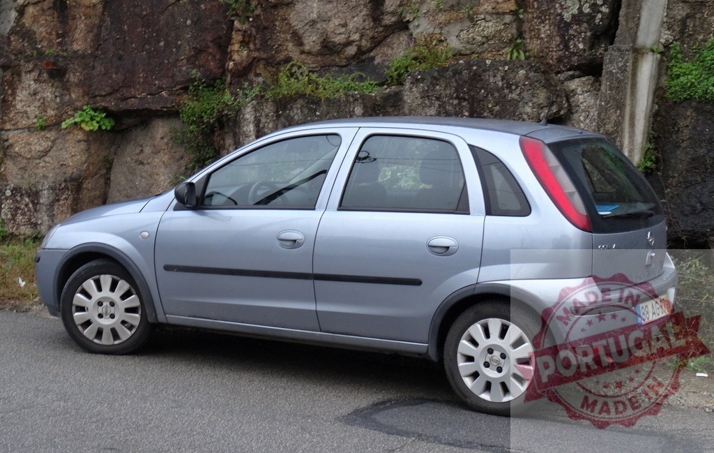 Opel Corsa parked on a road beside rock wall | Light blue compact five-door Opel Corsa parked by a natural rock wall on a quiet street - Realized with Pictrs.com