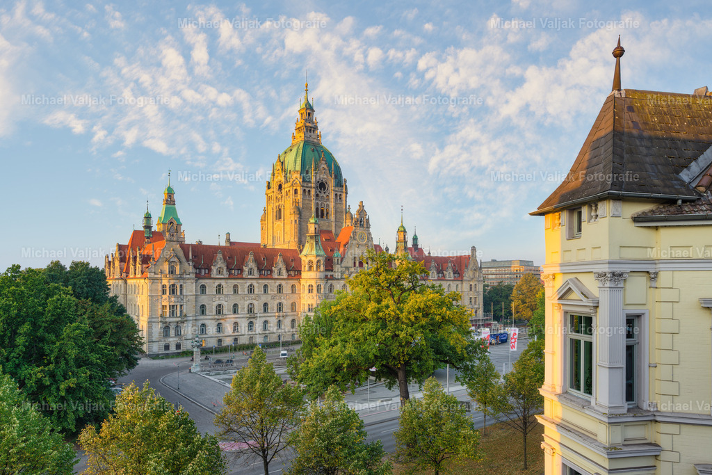 Neues Rathaus Hannover am Morgen | Das Neue Rathaus erscheint auf diesem Foto aus schräger Perspektive, von der frühen Morgensonne in warmes Licht getaucht. Die Fassade leuchtet in goldenen Tönen, während ein Teil eines anderen Gebäudes im Vordergrund den Blick rahmt und einen spannenden Kontrast setzt – stilistisch wie farblich. Diese Komposition lenkt den Fokus auf die monumentale Präsenz des Rathauses und betont zugleich die Vielfalt der hannoverschen Stadtarchitektur. - Realisiert mit Pictrs.com