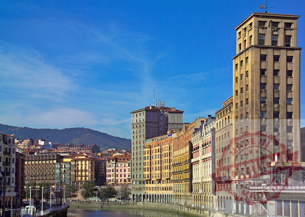 Bilbao city skyline along Nervion River waterfront in Basque Country. Spain - Bilbao | Bilbao city buildings lining Nervion River with mountains under blue sky - Realized with Pictrs.com