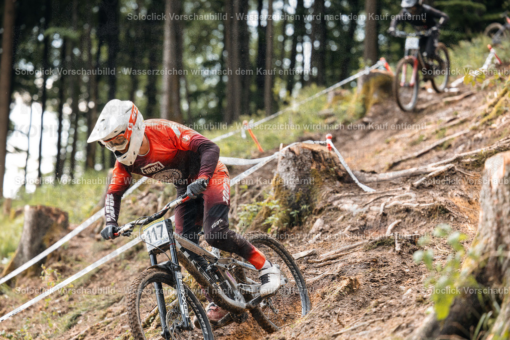 Mountainbiker im Full Speed | Ein dynamisches Foto eines Mountainbikers, der einen steilen, bewaldeten Hang hinunterfährt. Der Fokus liegt auf dem Fahrer, der mit hoher Geschwindigkeit einen Sprung vorbereitet. Die Umgebung besteht aus dichtem Wald und einem unebenen Gelände mit markierten Abschnitten. - Realisiert mit Pictrs.com